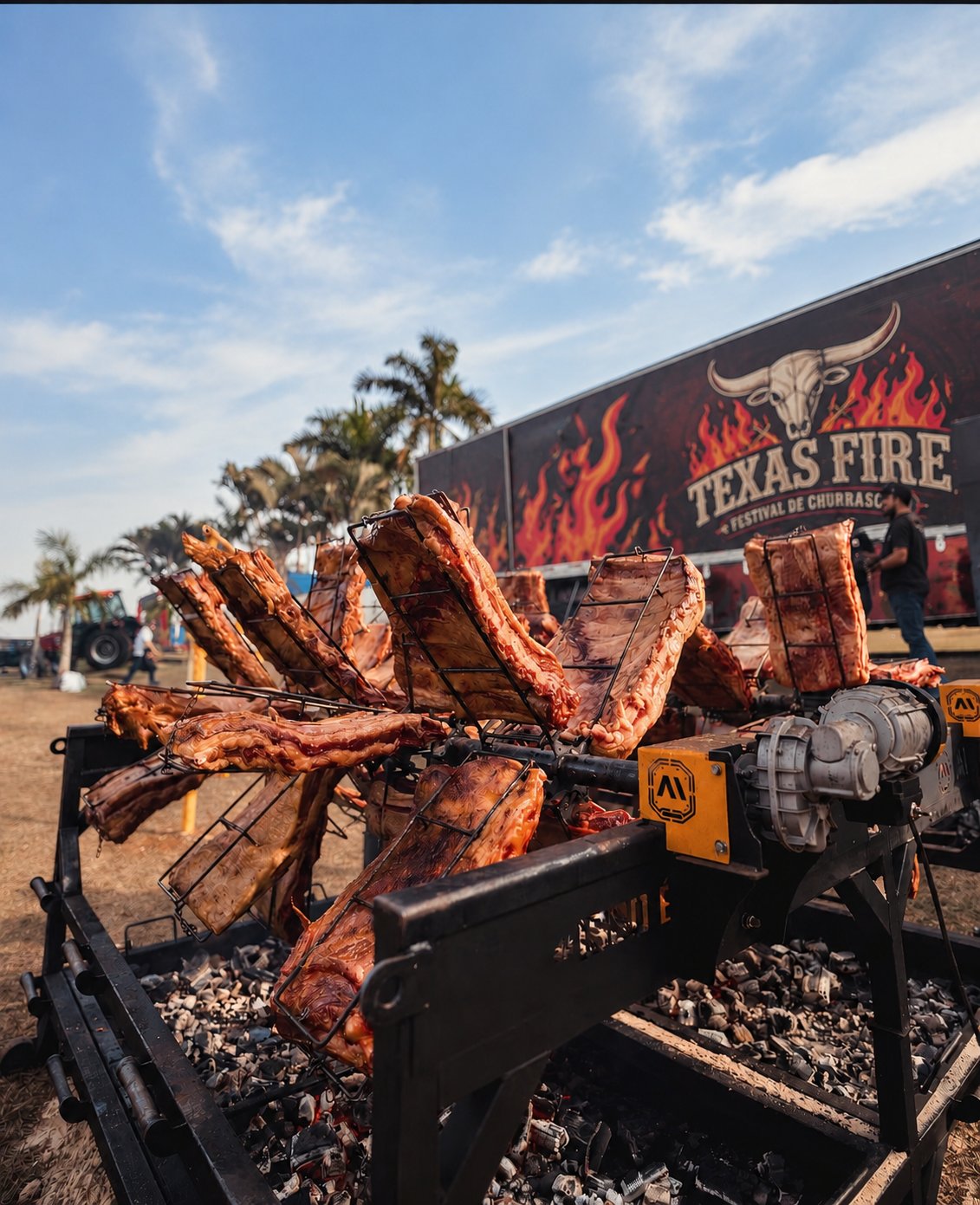 Estação de churrasco com costelas em grelhas giratórias sobre brasa, com o painel do Texas Fire ao fundo